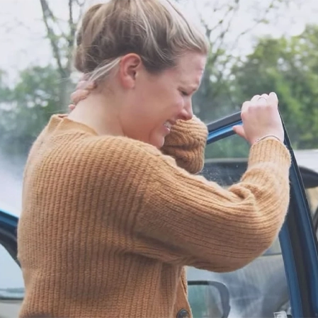 A person in a brown knitted sweater is standing next to an open car door, holding the door frame with one hand. Trees and a vehicle are visible in the background.