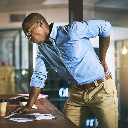 Person leaning on a desk, holding their lower back, indicating back pain.